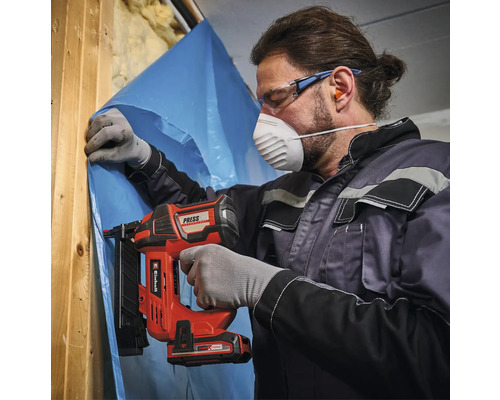 Mann befestigt mit einem Akku-Nagler eine Folie an einer Holzkonstruktion. Er trägt dabei eine Schutzmaske und Schutzbrille.