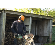 Un homme scie du bois avec une tronçonneuse électrique devant une remise à bois remplie de bûches.