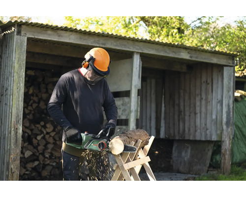 Un homme scie du bois avec une tronçonneuse électrique devant une remise à bois remplie de bûches.