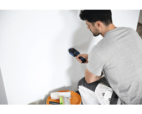 Un homme utilise un détecteur sur un mur près d'un seau contenant des outils de peintre