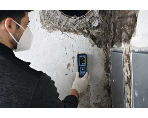 Eine Person mit Schutzmaske und Handschuhen führt mit einem Feuchtigkeitsmessgerät eine Feuchtigkeitsmessung an einer Wand durch.