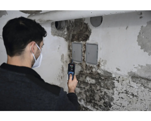 Eine Person verwendet ein Feuchtigkeitsmessgerät an einer beschädigten Wand in einem Gebäude.