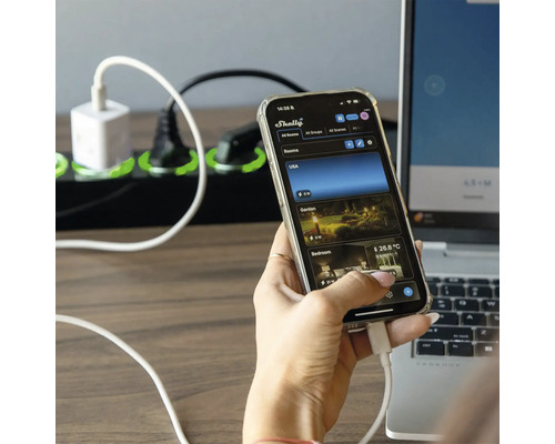 Eine Person bedient eine Smartphone App mit dem Shelly Logo, im Hintergrund eine Steckdosenleiste und ein Laptop