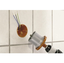 Scie cloche diamantée perçant un trou dans un mur carrelé à côté d'une boîte de dérivation avec des câbles électriques.