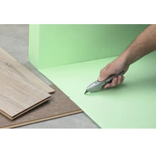 Une personne coupe une sous-couche isolante verte avec un cutter, à côté de laquelle se trouve du stratifié.