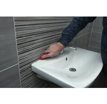 Une personne retire les joints en silicone entre le lavabo et le carrelage mural avec un outil.