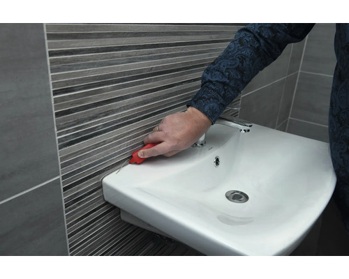 Une personne retire les joints en silicone entre le lavabo et le carrelage mural avec un outil.