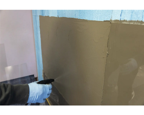 Eine Person befeuchtet eine Wand mit einer Sprühflasche, auf der bereits Putz aufgetragen wurde.