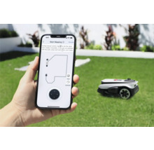 Une personne tient un smartphone avec une application pour contrôler un robot tondeuse dans le jardin.