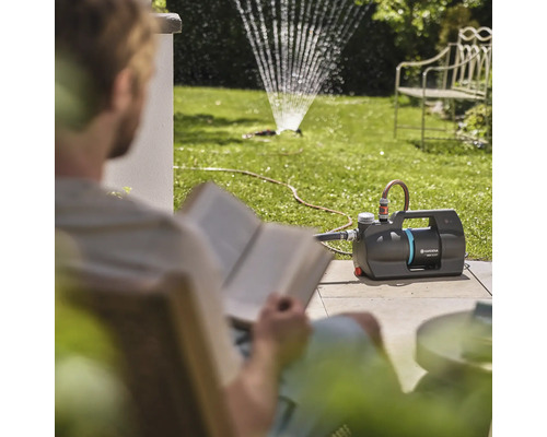Garten mit Rasensprenger und lesendem Mann neben einer Gartenpumpe