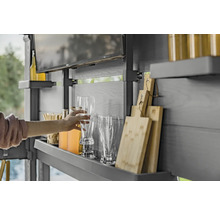 Eine Person greift nach einem Bierglas in einem Outdoor-Küchenbereich mit Regalen, Getränken, Gläsern, Schneidebrettern und einem Fernseher.