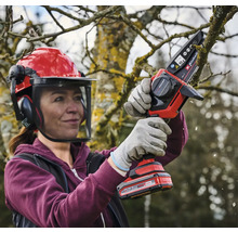 Une femme portant un casque de sécurité et des gants utilise une tronçonneuse sans fil pour couper une branche.