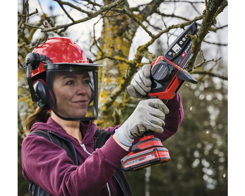 Une femme portant un casque de sécurité et des gants utilise une tronçonneuse sans fil pour couper une branche.