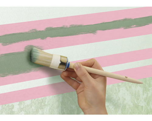 Une personne peint une ligne verte avec un pinceau rond sur un mur avec des rayures roses et blanches