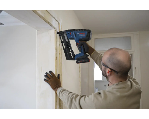 Un artisan fixe une latte sur un encadrement de porte à l'aide d'un cloueur à batterie.