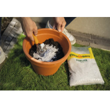 Gartenszene mit Person, die Perlite mit einer Schaufel in einen Pflanztopf mit Erde mischt, und eine Packung Flora Gard Perlite.