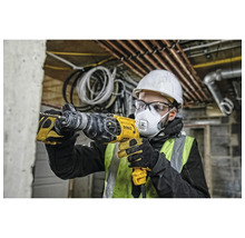 Un homme avec un casque de protection, des lunettes de protection et un masque respiratoire perce un mur avec une perceuse jaune.