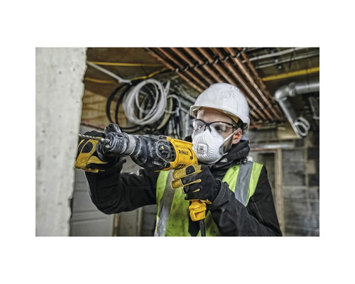 Un homme avec un casque de protection, des lunettes de protection et un masque respiratoire perce un mur avec une perceuse jaune.