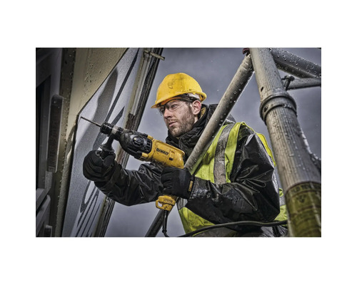 Un ouvrier perce un trou dans un mur avec une perceuse, debout sur un échafaudage.