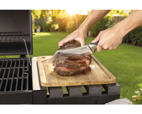 Un steak cuit est coupé au couteau sur une planche à découper en bois à côté d'un barbecue.
