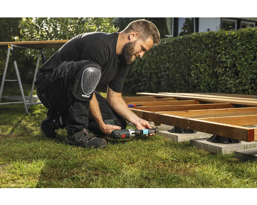 Ein Mann baut eine Holzterrasse mit einem Akku-Bohrschrauber und Knieschützern im Garten.