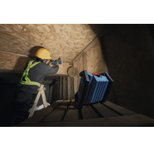 Un artisan portant un casque de protection perce un plafond en bois avec une visseuse sans fil, tandis qu'une lampe de travail se trouve au premier plan.