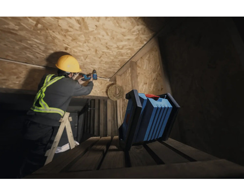 Un artisan portant un casque de protection perce un plafond en bois avec une visseuse sans fil, tandis qu'une lampe de travail se trouve au premier plan.