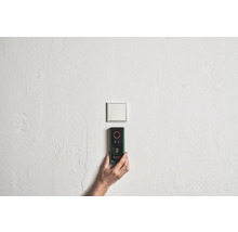 Une main tient un télémètre Bosch sur un mur près d'un interrupteur.