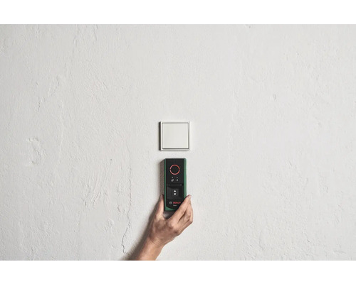 Une main tient un télémètre Bosch sur un mur près d'un interrupteur.