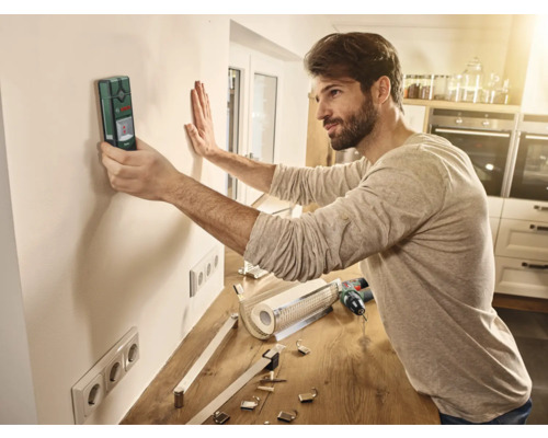 Un homme utilise un détecteur sur un mur dans une pièce pour préparer des travaux d'installation.