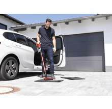 Un homme aspire un tapis de sol devant une voiture avec un aspirateur sans fil.