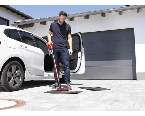 Un homme aspire un tapis de sol devant une voiture avec un aspirateur sans fil.