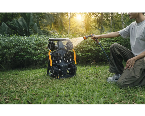 Un homme nettoie un robot tondeuse avec un tuyau d'arrosage dans le jardin.