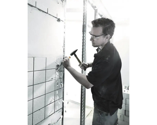 Mann mit Schutzbrille und Hammer bei Renovierungsarbeiten an einer gefliesten Wand.