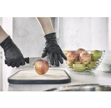 Une personne portant des gants de protection coupe une pomme sur une planche à découper avec un couteau, avec un bol de morceaux de pomme en arrière-plan.