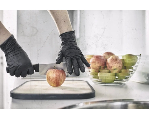 Une personne portant des gants de protection coupe une pomme sur une planche à découper avec un couteau, avec un bol de morceaux de pomme en arrière-plan.