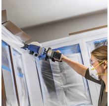 Femme peignant un cadre de fenêtre avec un pulvérisateur de peinture, les fenêtres sont recouvertes d'un film plastique