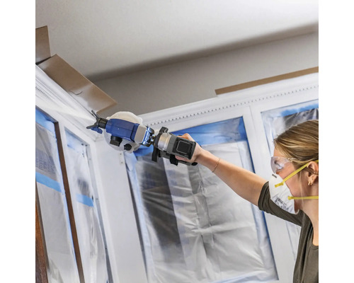 Femme peignant un cadre de fenêtre avec un pulvérisateur de peinture, les fenêtres sont recouvertes d'un film plastique