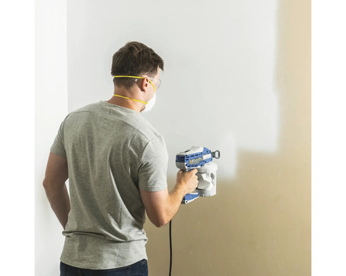 Mann streicht Wand mit Farbsprühgerät, ausgestattet mit Schutzbrille und Staubmaske