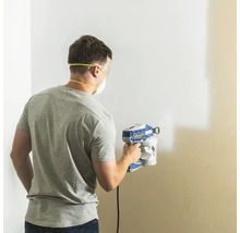 Homme peignant un mur avec un pulvérisateur de peinture, équipé de lunettes de protection et d'un masque anti-poussière