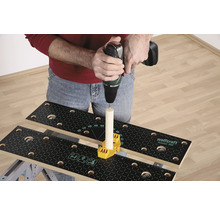 Eine Person bohrt mit einer Bohrmaschine in ein Holzstück, das auf einer Werkbank mit gelbem Befestigungselement befestigt ist.