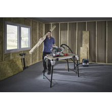 Mann mit Holzbalken, Kappsäge und Werkbank in einem Rohbau.