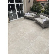 Moderne Terrasse mit hellgrauen quadratischen Terrassenplatten in Steinoptik, zwei grauen Gartensofas aus Geflecht und großen Fensterelementen.