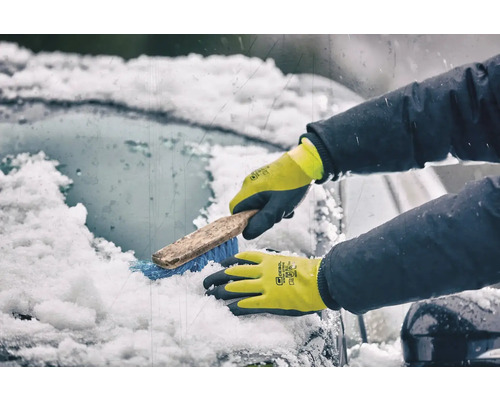 Une personne portant des gants d'hiver nettoie un pare-brise enneigé avec une brosse.
