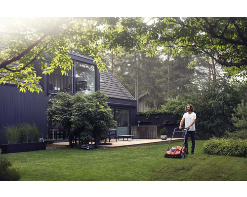 Mann mäht Rasen mit einem Rasenmäher in einem Garten vor einem modernen Haus.
