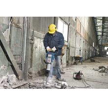 Un homme portant un casque de sécurité et une protection auditive utilise un marteau-piqueur à l'intérieur.