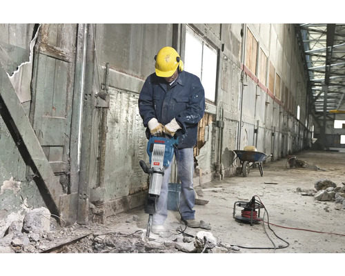 Un homme portant un casque de sécurité et une protection auditive utilise un marteau-piqueur à l'intérieur.