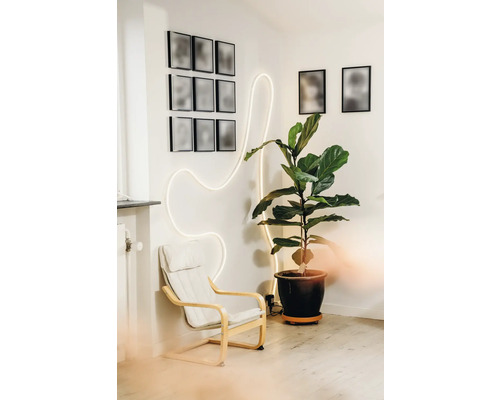 Salon moderne avec un fauteuil blanc, une grande plante verte en pot et un ruban lumineux LED ondulé sur un mur blanc.