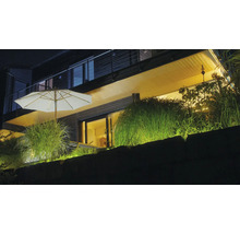 Terrasse moderne éclairée de nuit avec parasol blanc, graminées ornementales et éclairage extérieur chaleureux sur un mur en pierre.