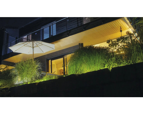 Terrasse moderne éclairée de nuit avec parasol blanc, graminées ornementales et éclairage extérieur chaleureux sur un mur en pierre.